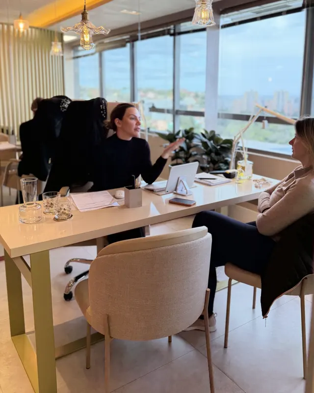 A Dra. Marina conversando com uma paciente na mesa do consultório, com janelas para a cidade