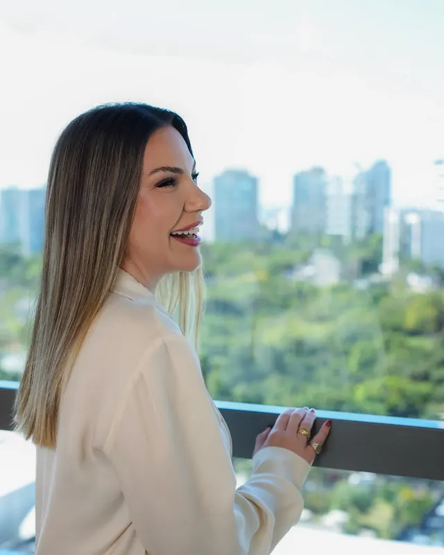 A Dra. Marina sorrindo diante de uma janela com vista para o verde da cidade